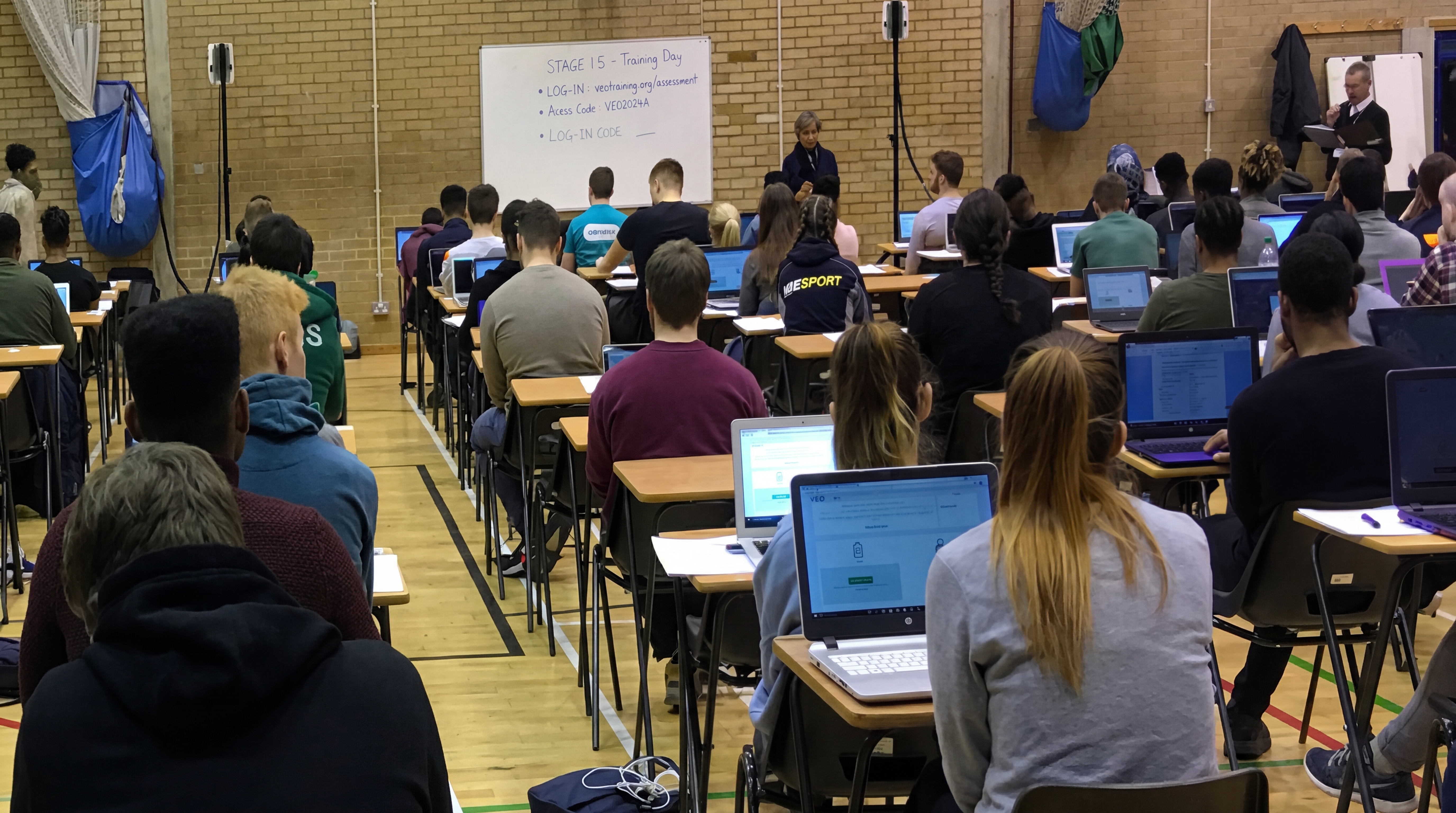 Students taking a digital exam on laptops in a school hall